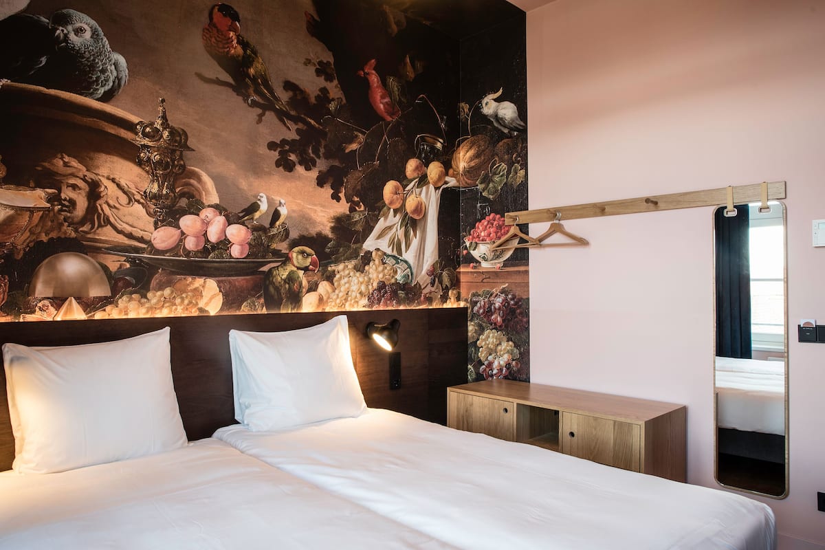 A bedroom with a large bed, a wooden headboard, a wall-mounted mirror, and a mural of fruits and birds.