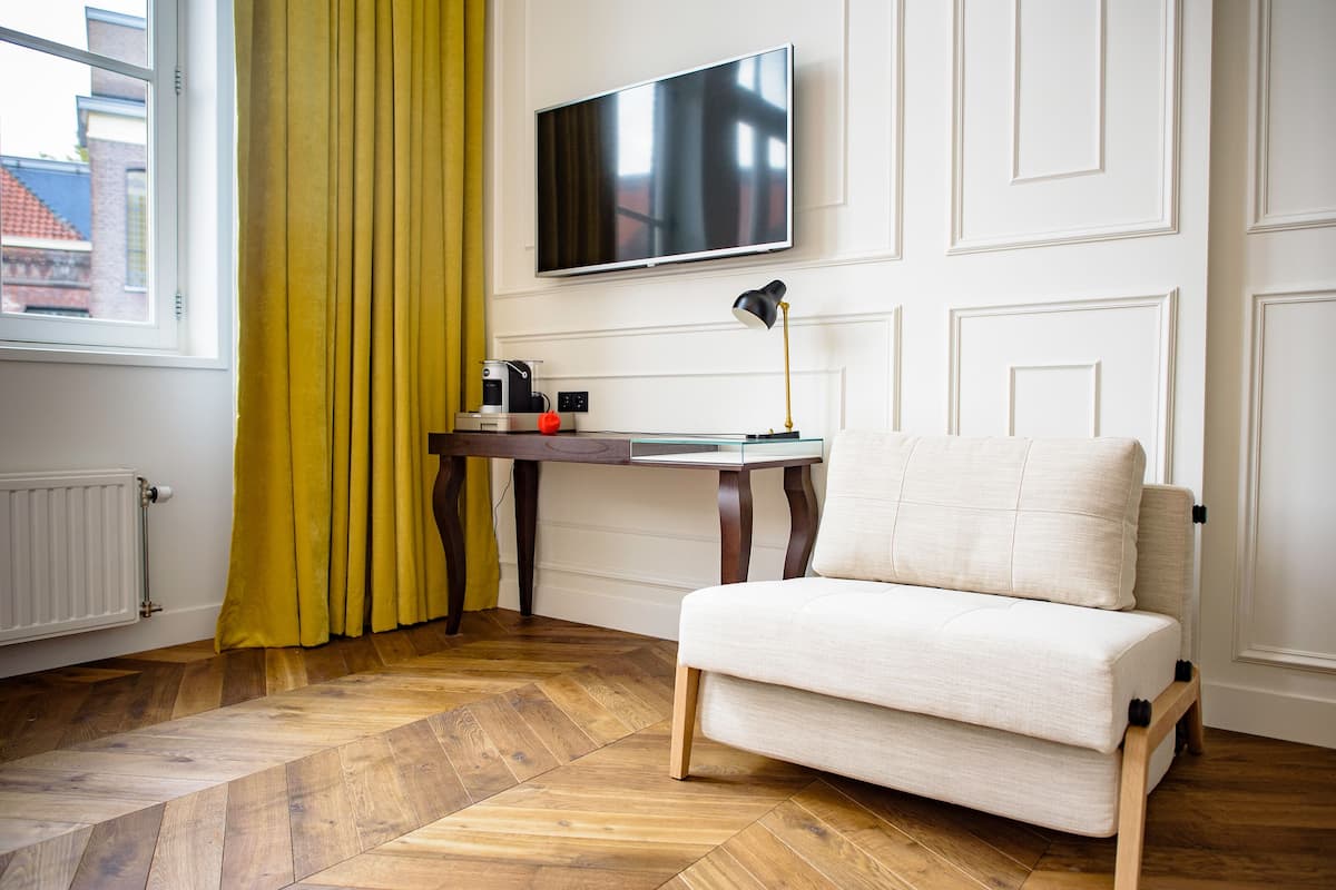 A room with a sofa, a TV mounted on the wall, a table with a lamp, and a window with curtains.