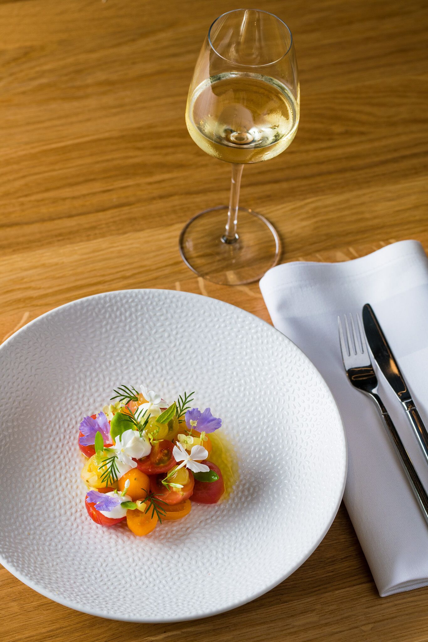 A white textured plate with a colorful salad, a glass of white wine, and cutlery on a wooden table.