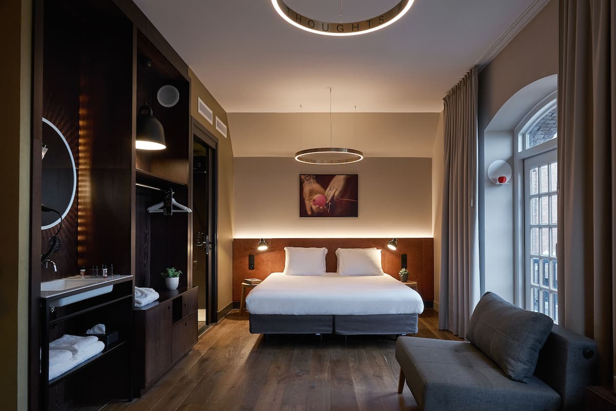 A modern hotel room with a large bed, a built-in wardrobe, a vanity with a mirror, and a large window with curtains.