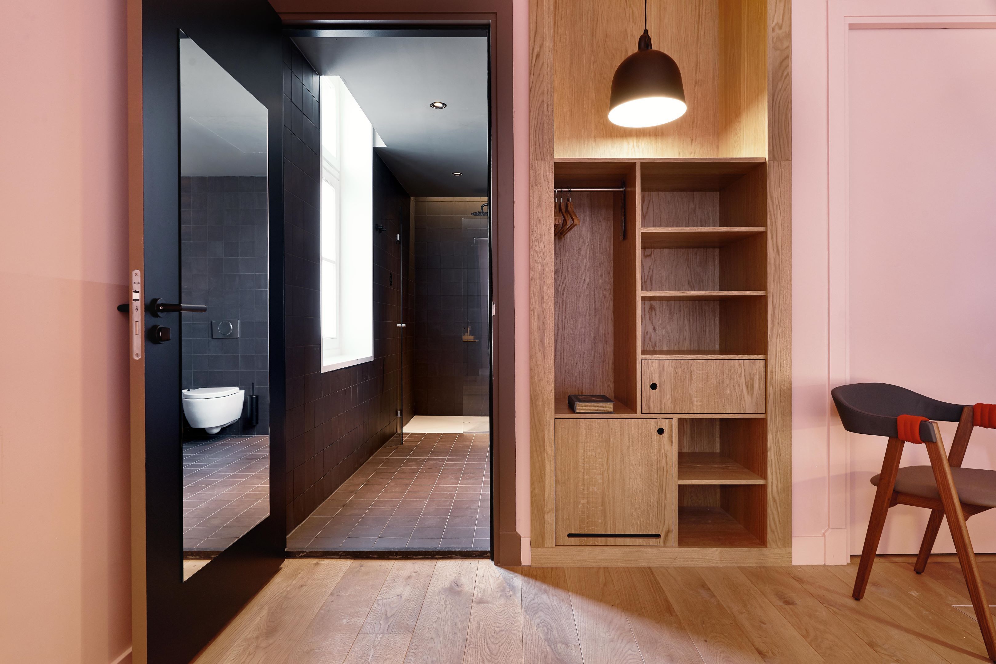 Un baño moderno con inodoro, lavabo y zona de ducha. Se observa un mueble de madera con estantes y un aplique colgante.