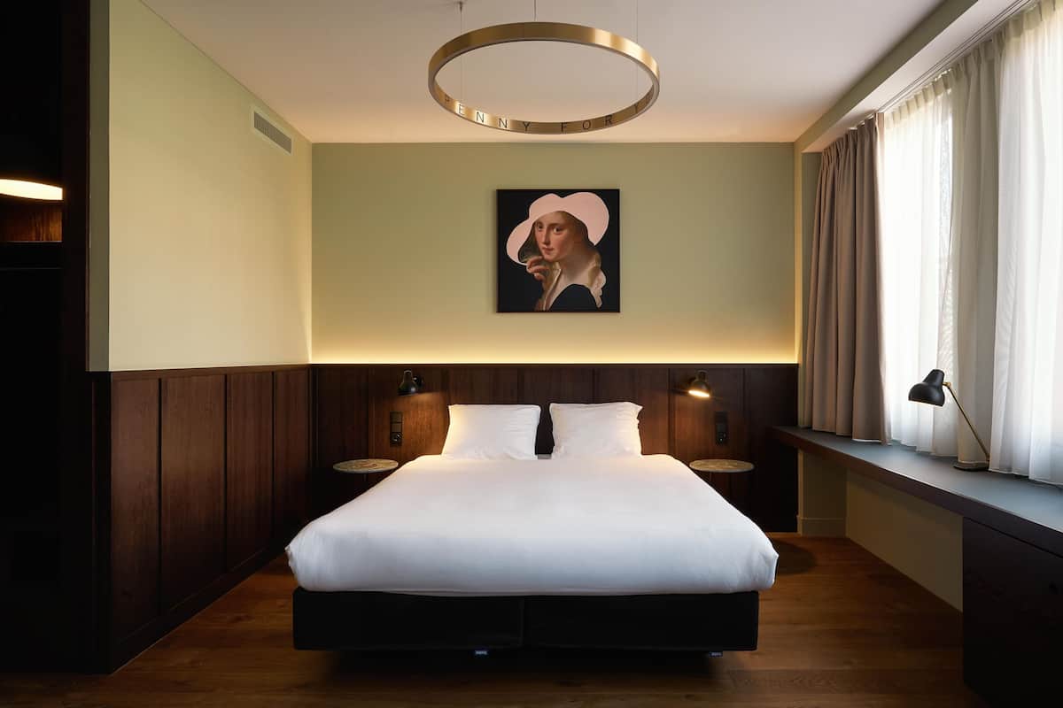 A modern hotel room with a large bed, wooden paneling, a circular light fixture, and a painting on the wall.