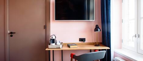 A room with a wooden desk, a chair, a flat-screen TV mounted on the wall, a coffee machine, a book, and a window with curtains.