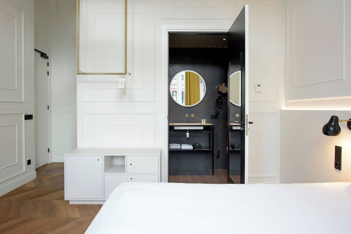 A modern hotel room with a large bed, a white wardrobe, a built-in vanity with a mirror, and a small seating area.