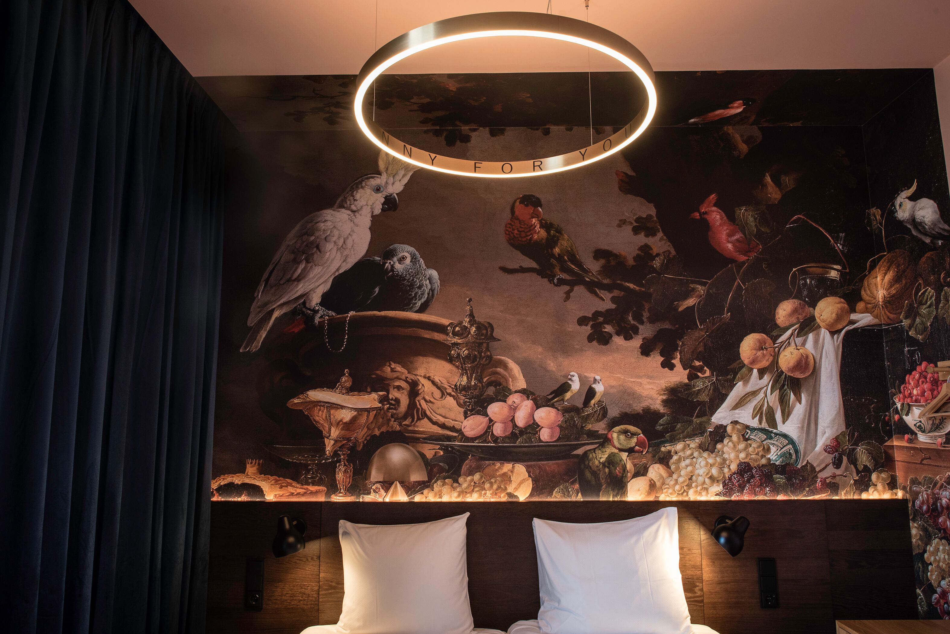 Una habitación con un mural de pájaros y frutas, una lámpara circular y dos almohadas blancas.