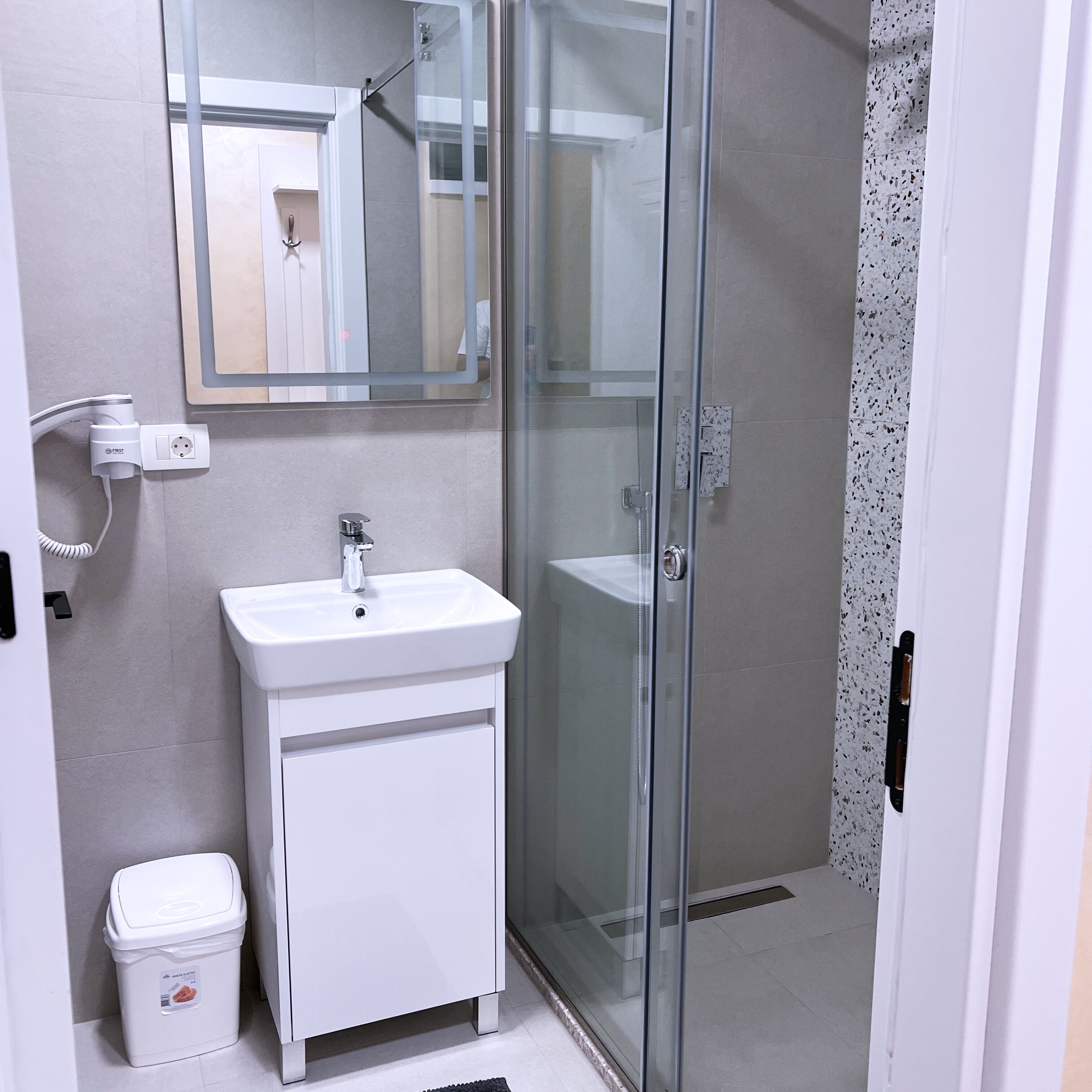 A modern bathroom with a glass shower enclosure, a white sink, and a trash bin.