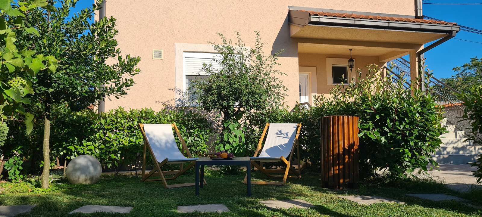 A garden with two lounge chairs, a table with fruit, and a building with a tiled roof and a window.