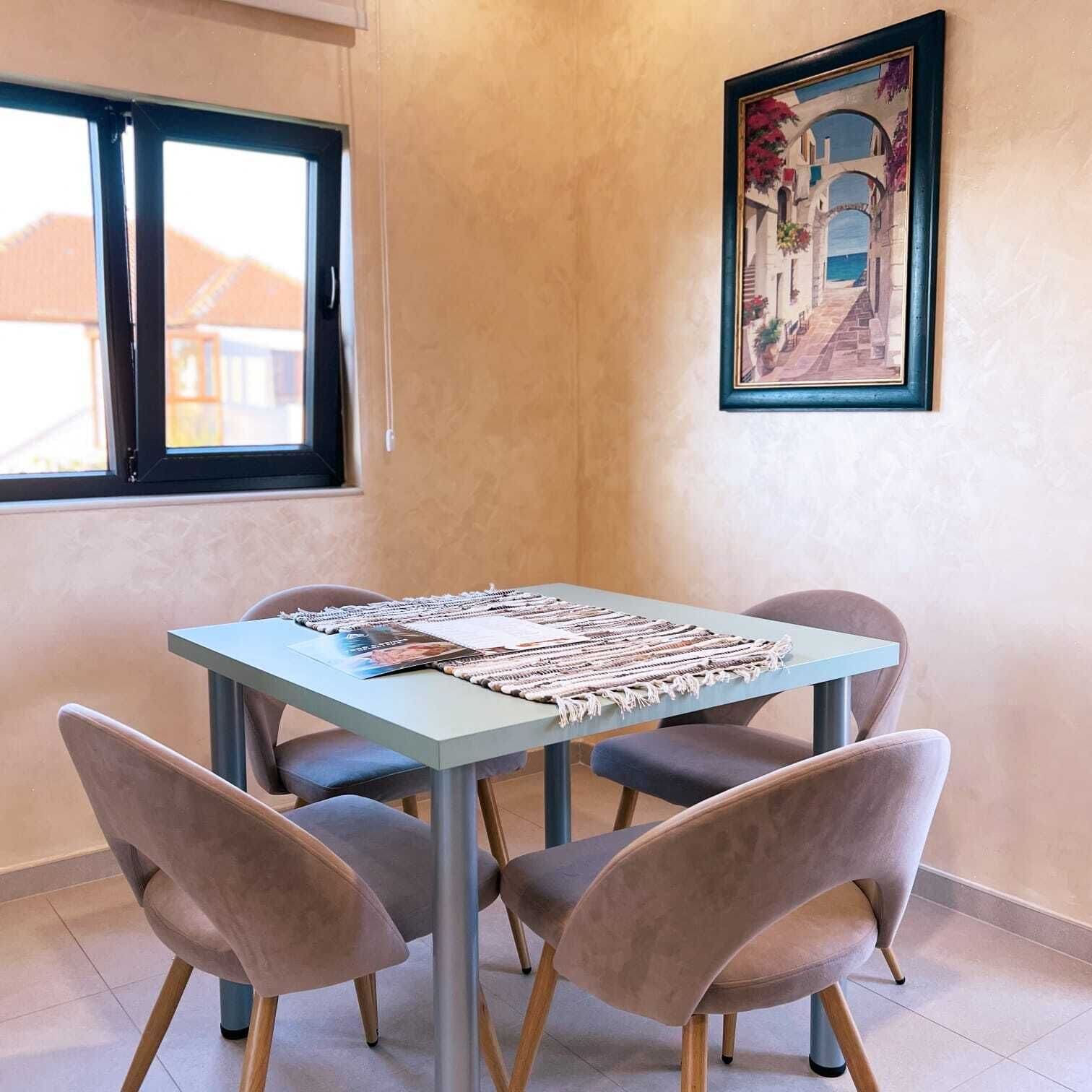 A dining area with a table, four chairs, a framed picture on the wall, and a window with blinds.