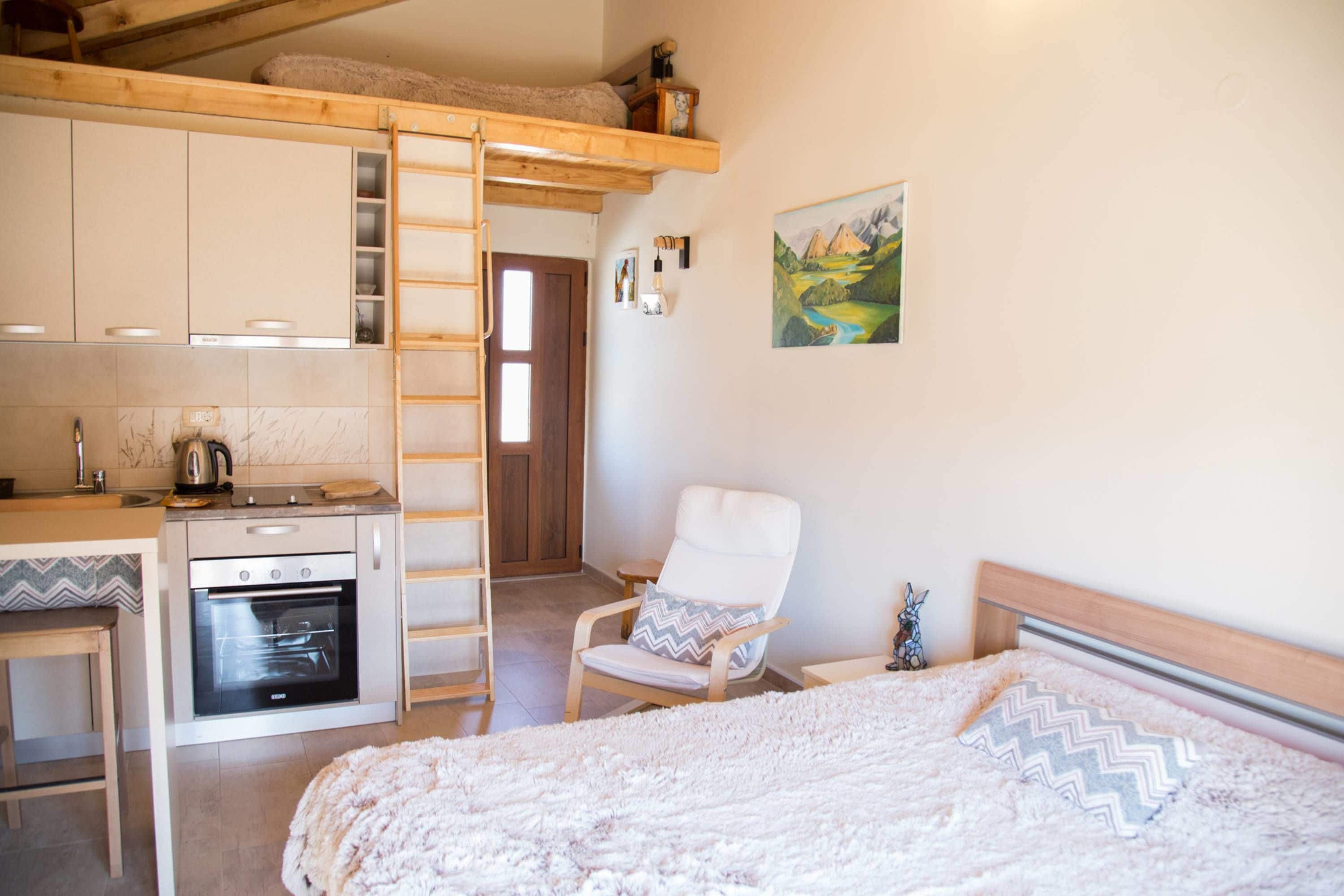 A compact room with a bunk bed, a kitchenette with an oven, a chair, and a painting on the wall.