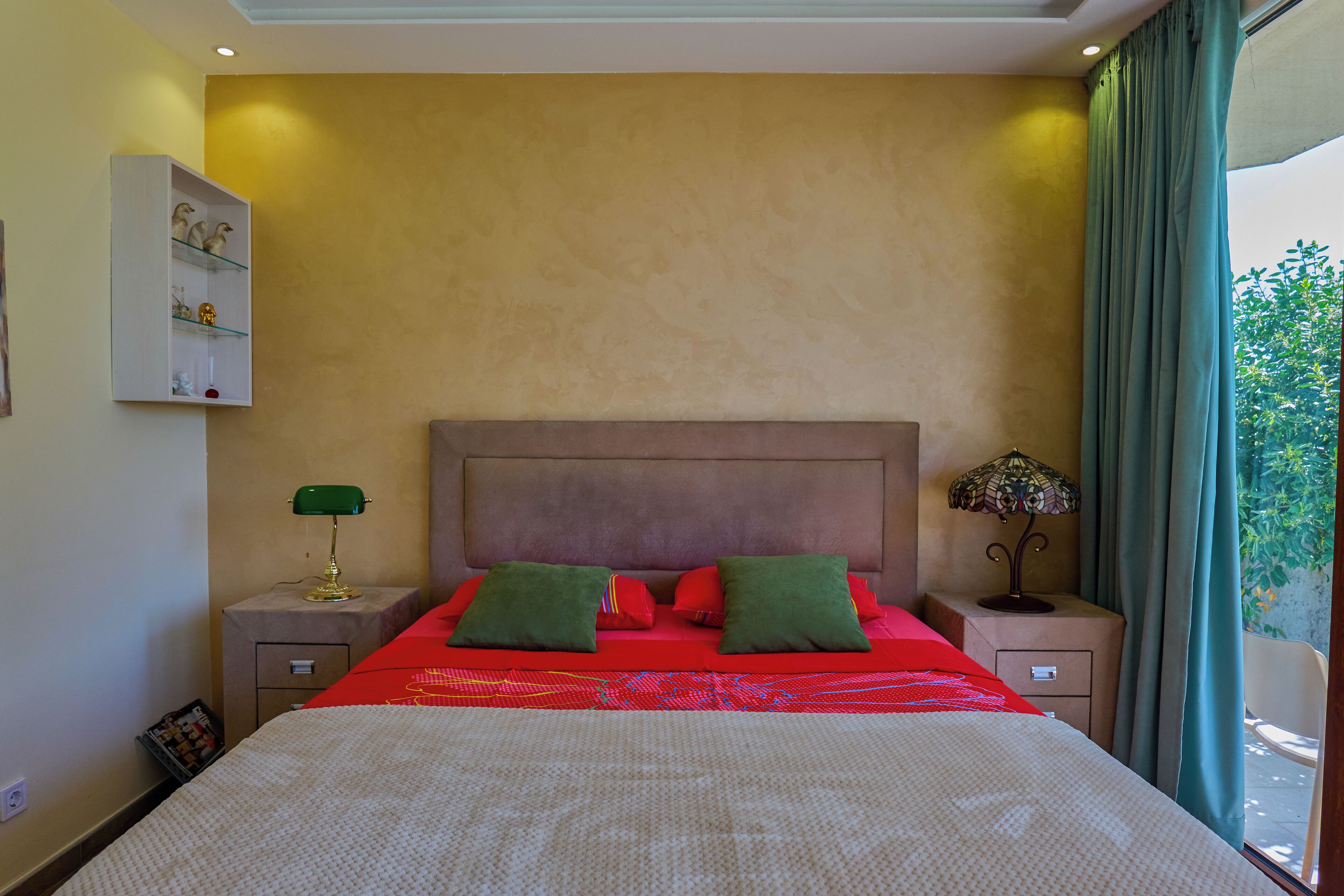 A bedroom with a large bed, a nightstand with a lamp, a dresser, and a view of greenery outside.
