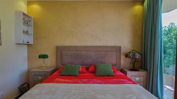 A bedroom with a large bed, a nightstand with a lamp, a dresser, and a view of greenery outside.