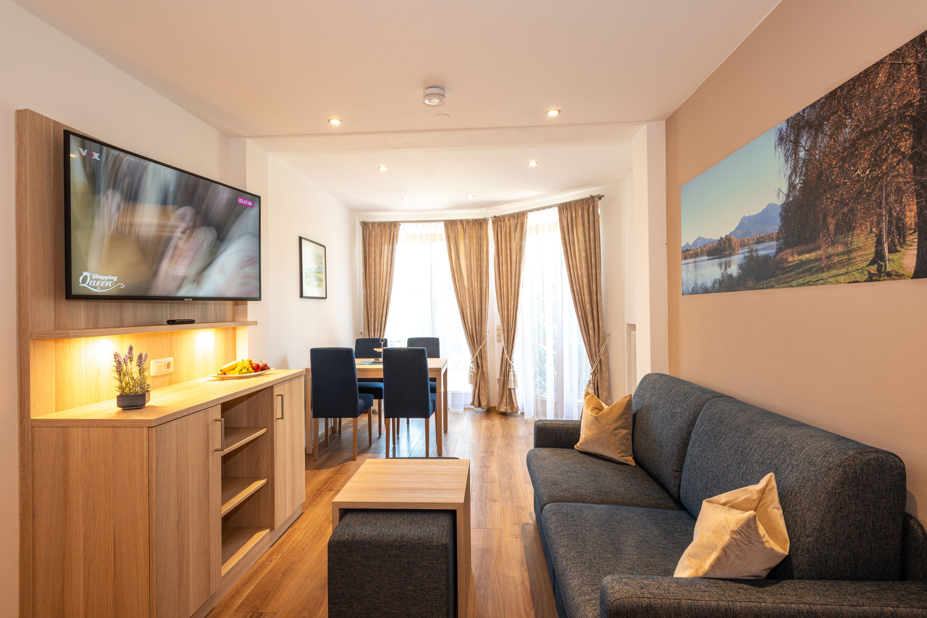 A modern living room with a grey sofa, a wooden dining table with chairs, a TV mounted on the wall, and a painting on the wall.