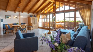 A spacious living area with a high ceiling, wooden beams, a large glass door leading to an outdoor patio, and a comfortable seating arrangement. - Aparthotel Werdenfels (Murnau am Staffelsee)