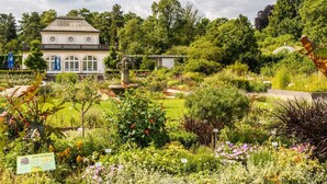 A garden with a variety of plants and flowers, a fountain, and a building with arched windows. - Aparthotel Werdenfels (Murnau am Staffelsee)