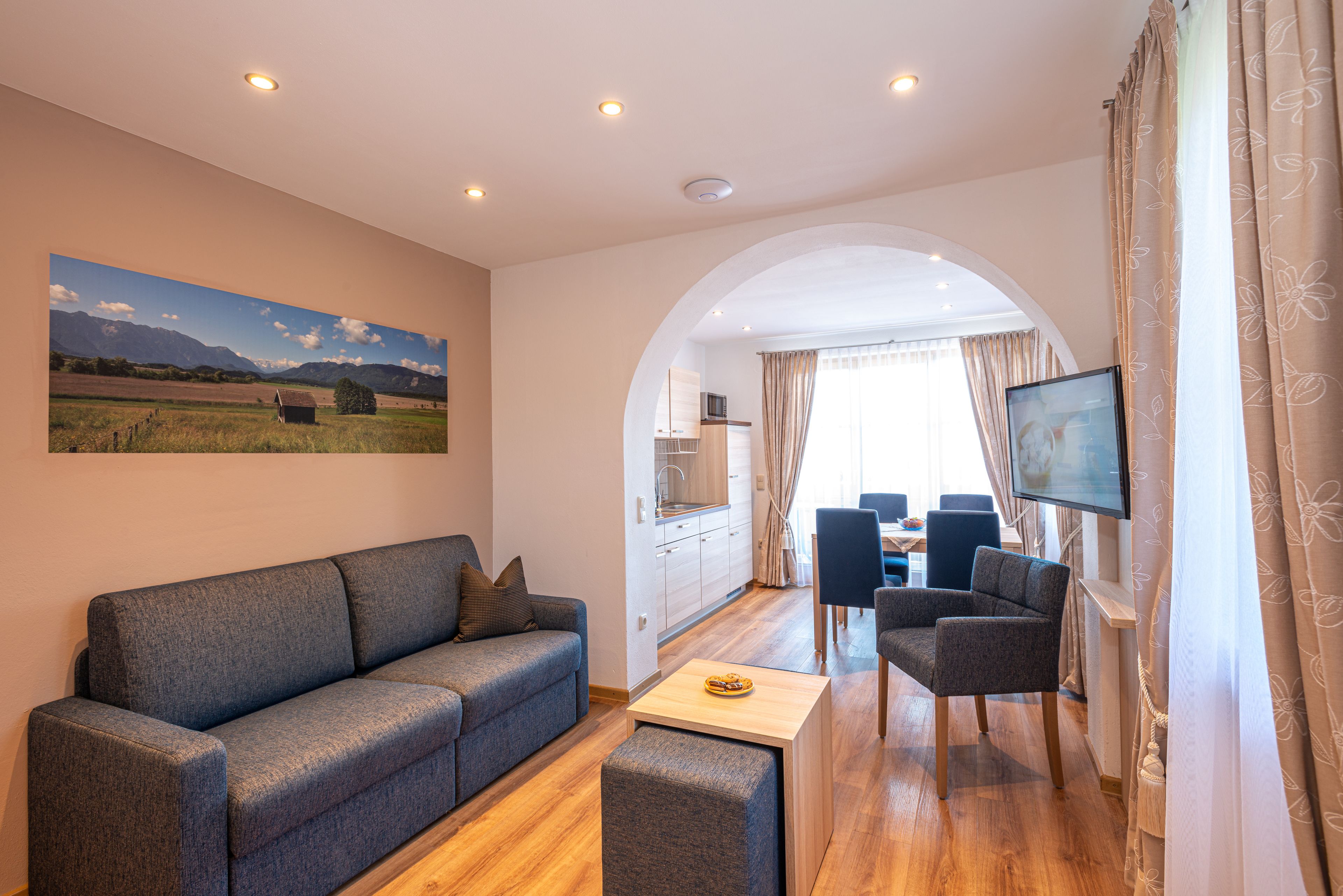 Una sala moderna con sofá, sillón y mesa de centro. Hay un televisor montado en la pared, un cuadro de un paisaje y un arco que da a un comedor.