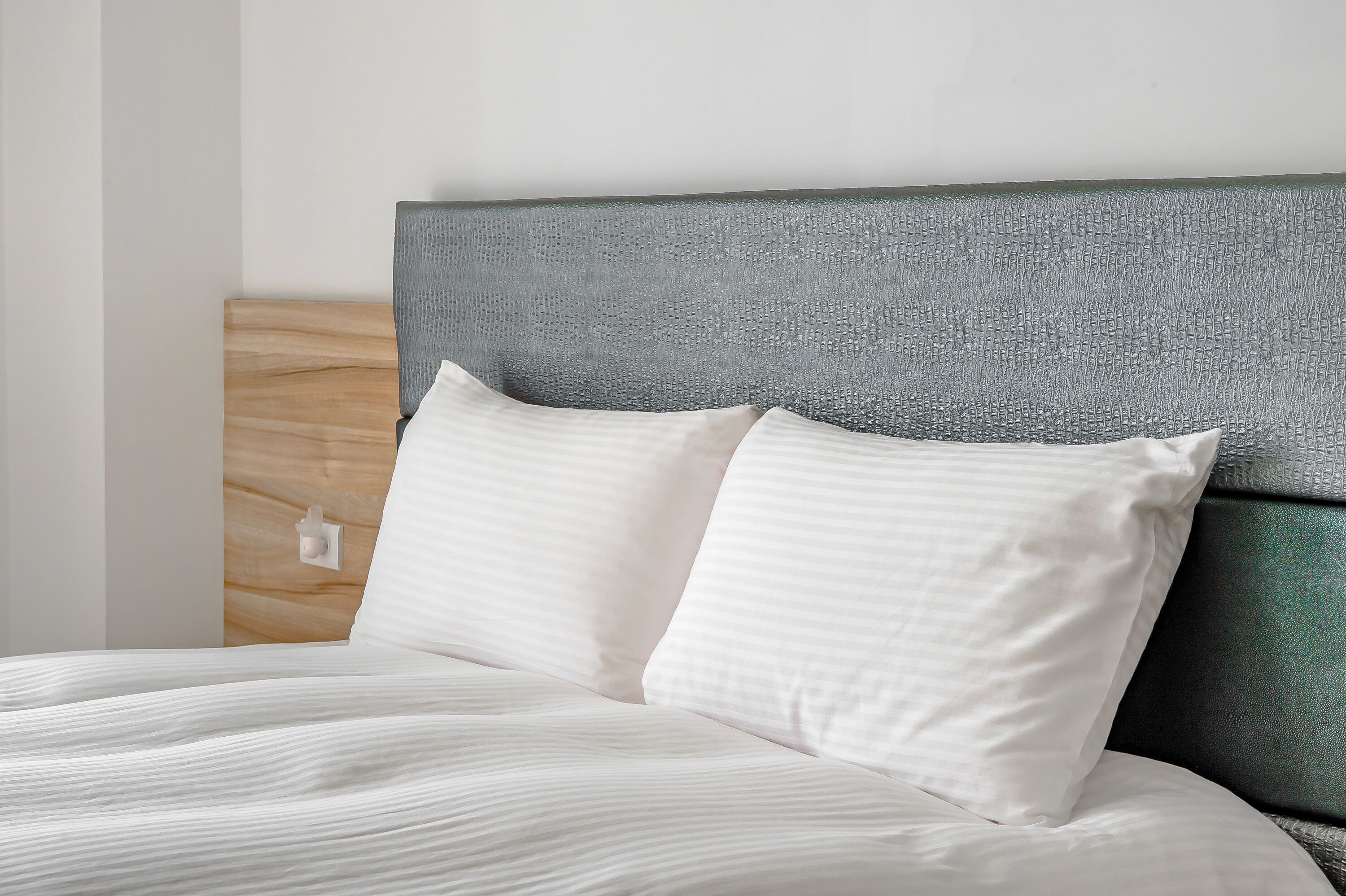 A neatly made bed with white linens and pillows, a wooden headboard, and a plain wall.