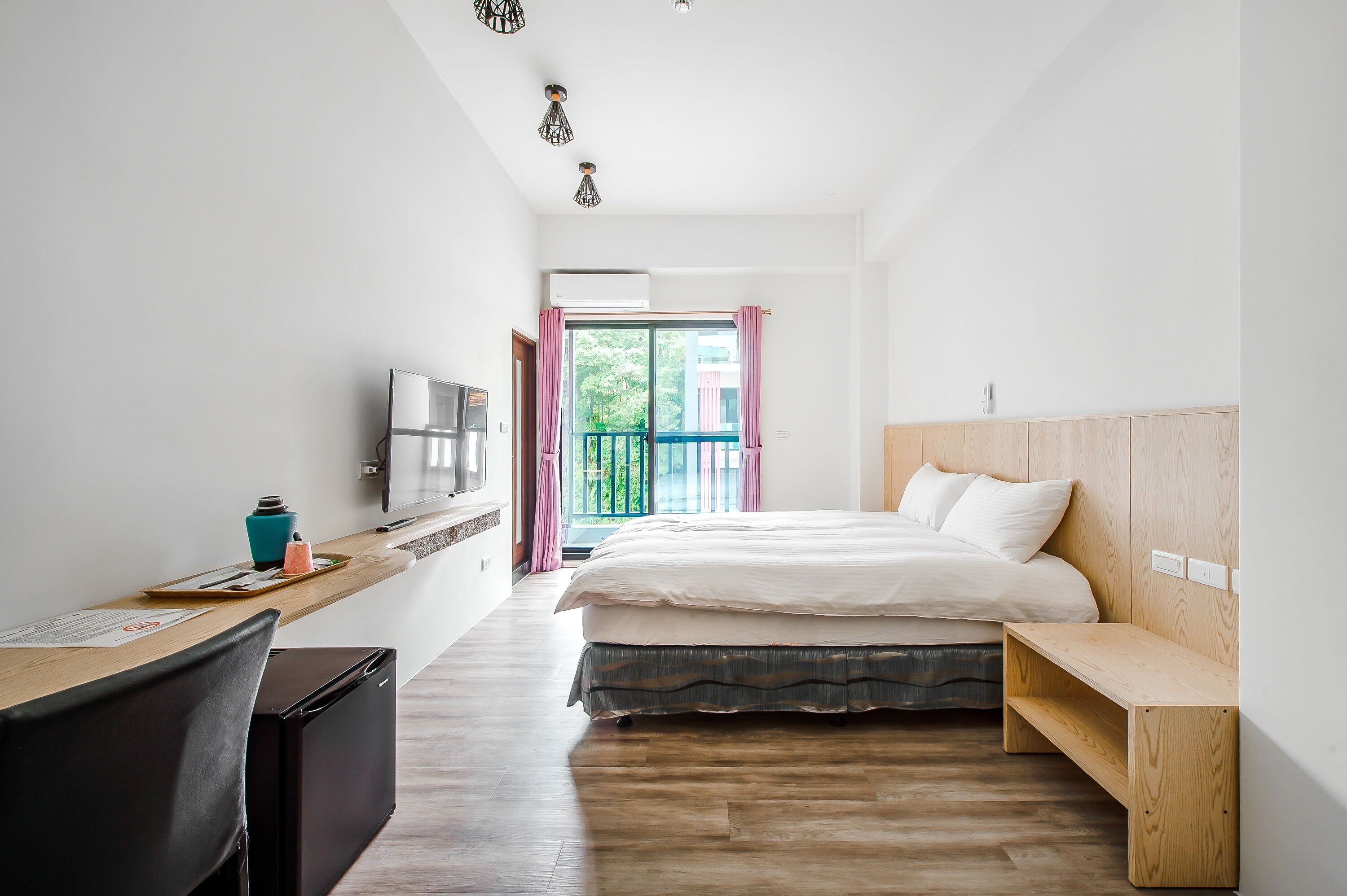 A modern hotel room with a wooden desk, a bed with a wooden headboard, a TV, and a balcony with a view of greenery.
