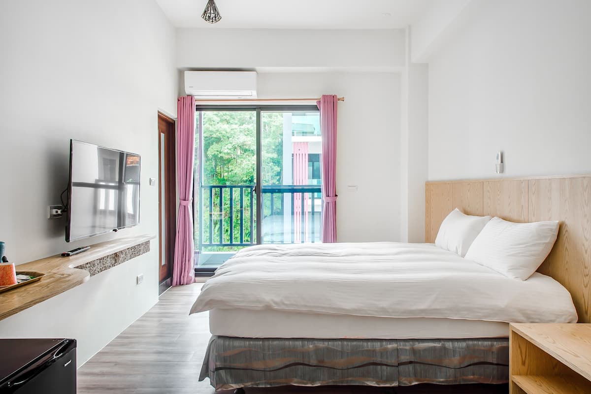 A bedroom with a bed, a TV, a wooden nightstand, and a balcony with pink curtains.