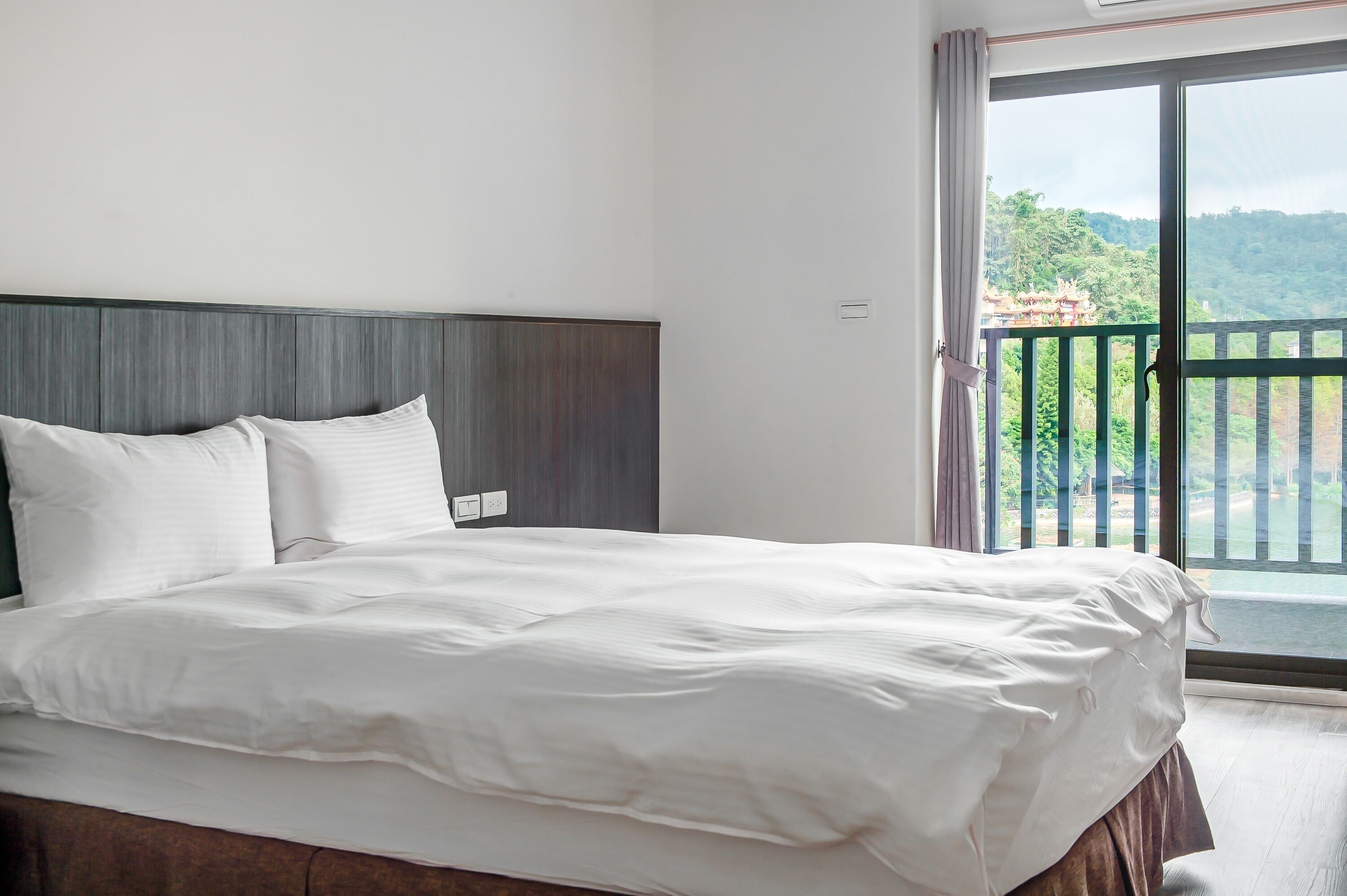 Habitación de hotel con una cama grande, cabecera de madera y un balcón con vistas a zonas verdes y un edificio.