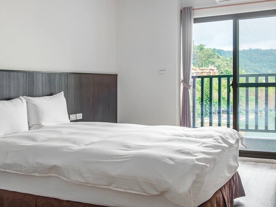 Habitación de hotel con una cama grande, cabecera de madera y un balcón con vistas a zonas verdes y un edificio.