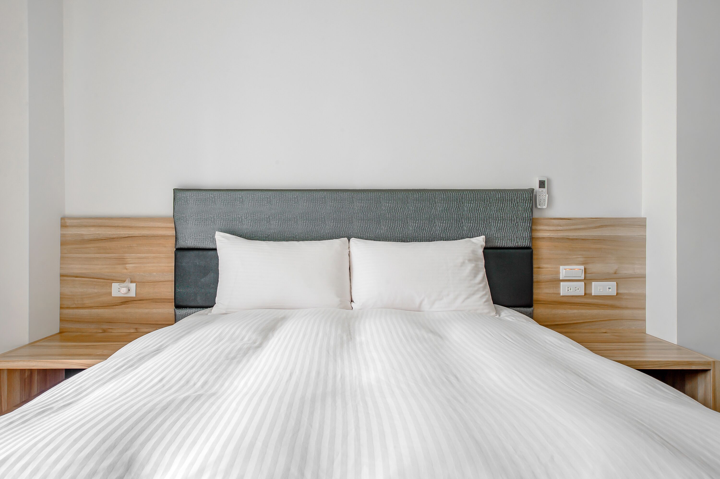 Una cama bien hecha con un edredón blanco a rayas, un cabecero de madera y dos mesitas de noche con un teléfono y un interruptor de luz.