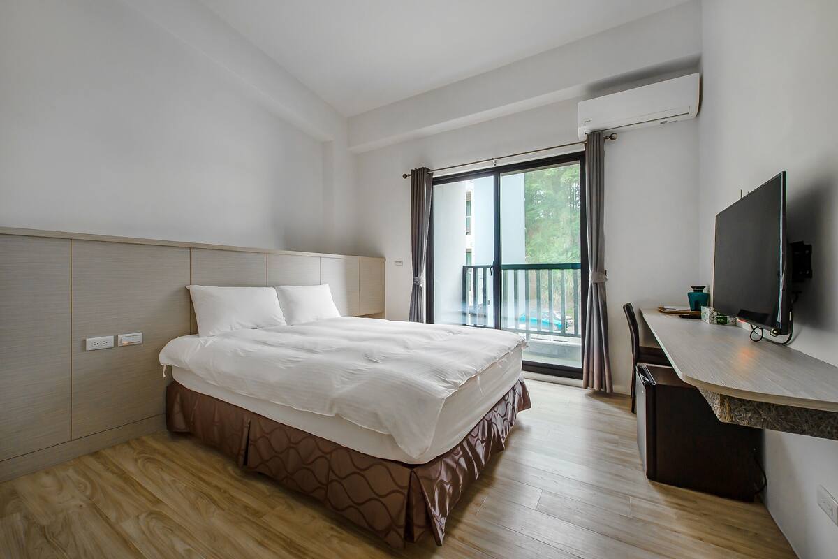 A hotel room with a bed, a desk, a TV, and a balcony with a view of greenery.