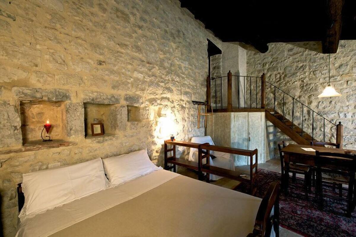 A rustic room with a stone wall, a wooden bed, a bench, a table, and a staircase.