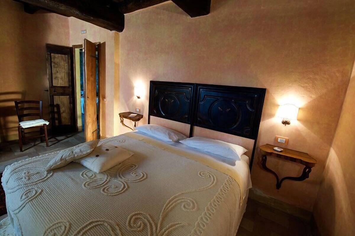 A bed with a headboard, bedside tables, and a chair in a room with a wooden ceiling and a door leading to another room.