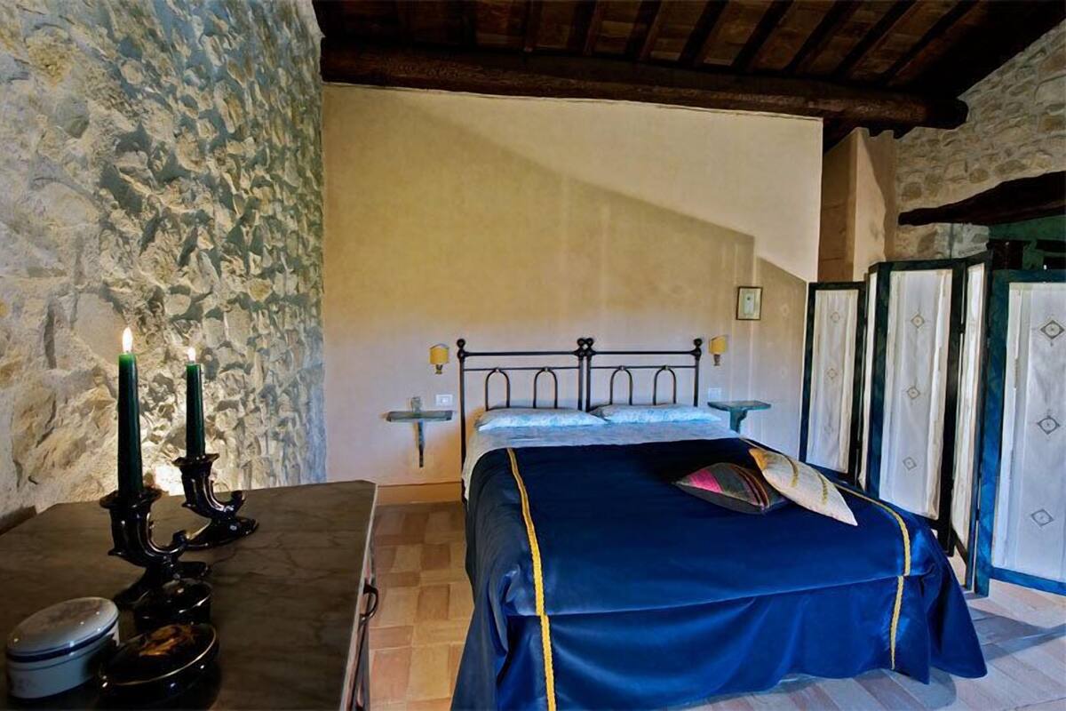 A bedroom with a stone wall, a bed with a blue bedspread, two lit candles on a table, and a small table with a candle holder.
