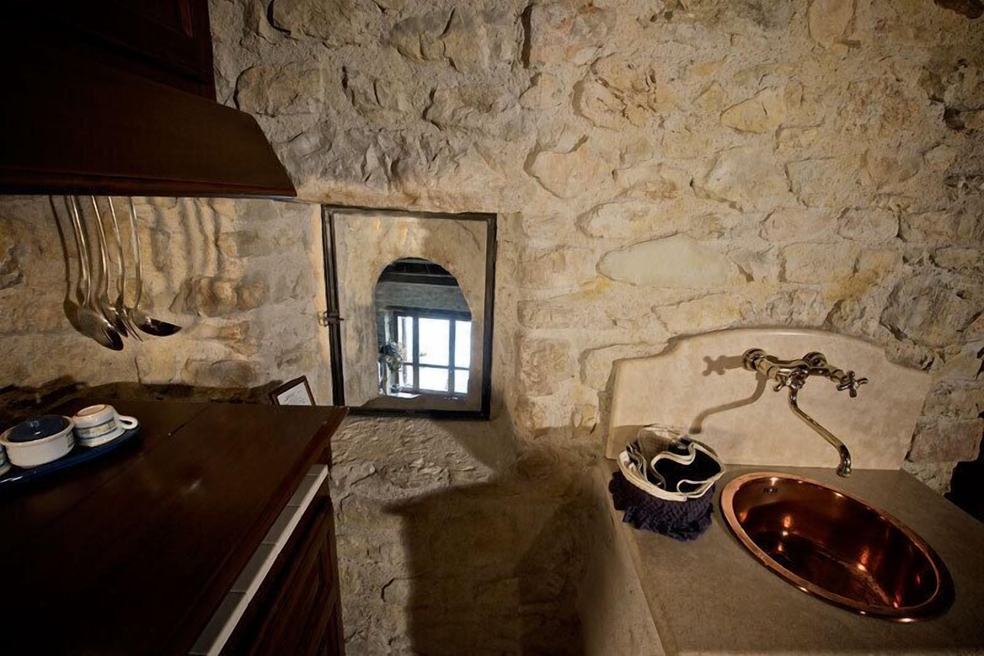 A rustic stone-walled room with a wooden table, a copper sink, and a small arched doorway leading to another room.