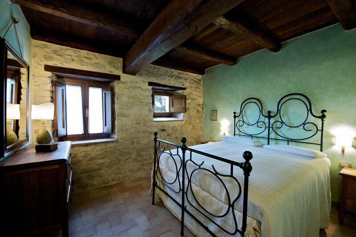 A bedroom with a stone wall, wooden ceiling beams, a metal bed frame, and a bedside table with a lamp.