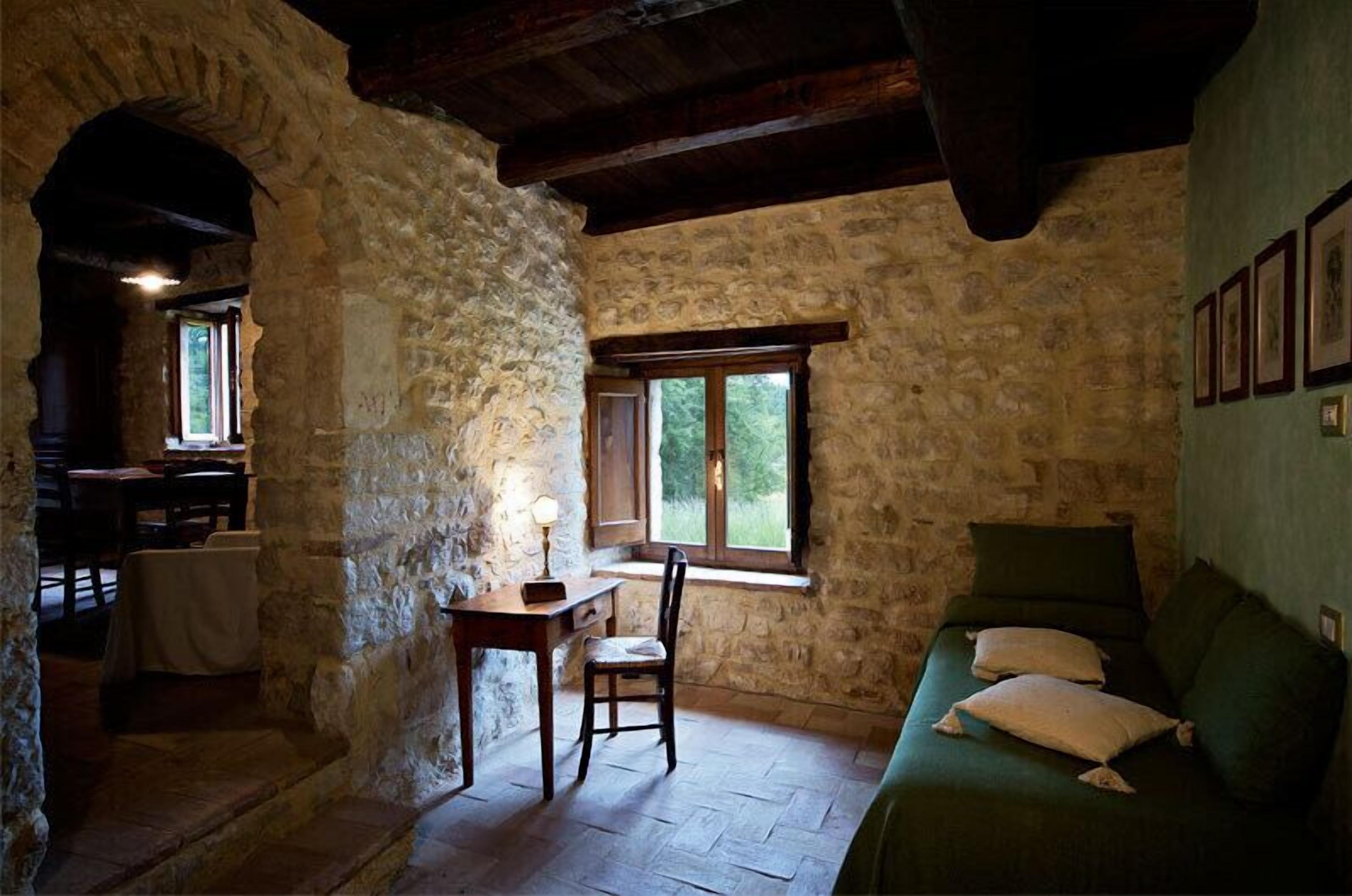 Ein rustikaler Raum mit Steinwänden, einem Holztisch mit Stuhl, einem Bett mit Kissen und einem Fenster mit Blick auf Grün.