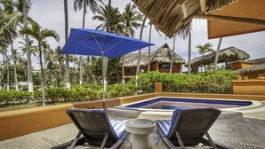 Un área junto a la piscina con sillones de descanso y una sombrilla azul, rodeada de palmeras y cabañas con techo de paja.