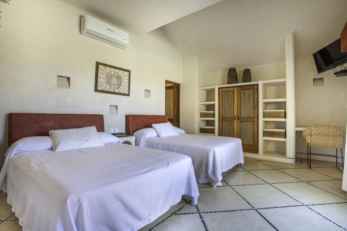 Habitación de hotel con dos camas, un televisor montado en la pared, una estantería y una silla.