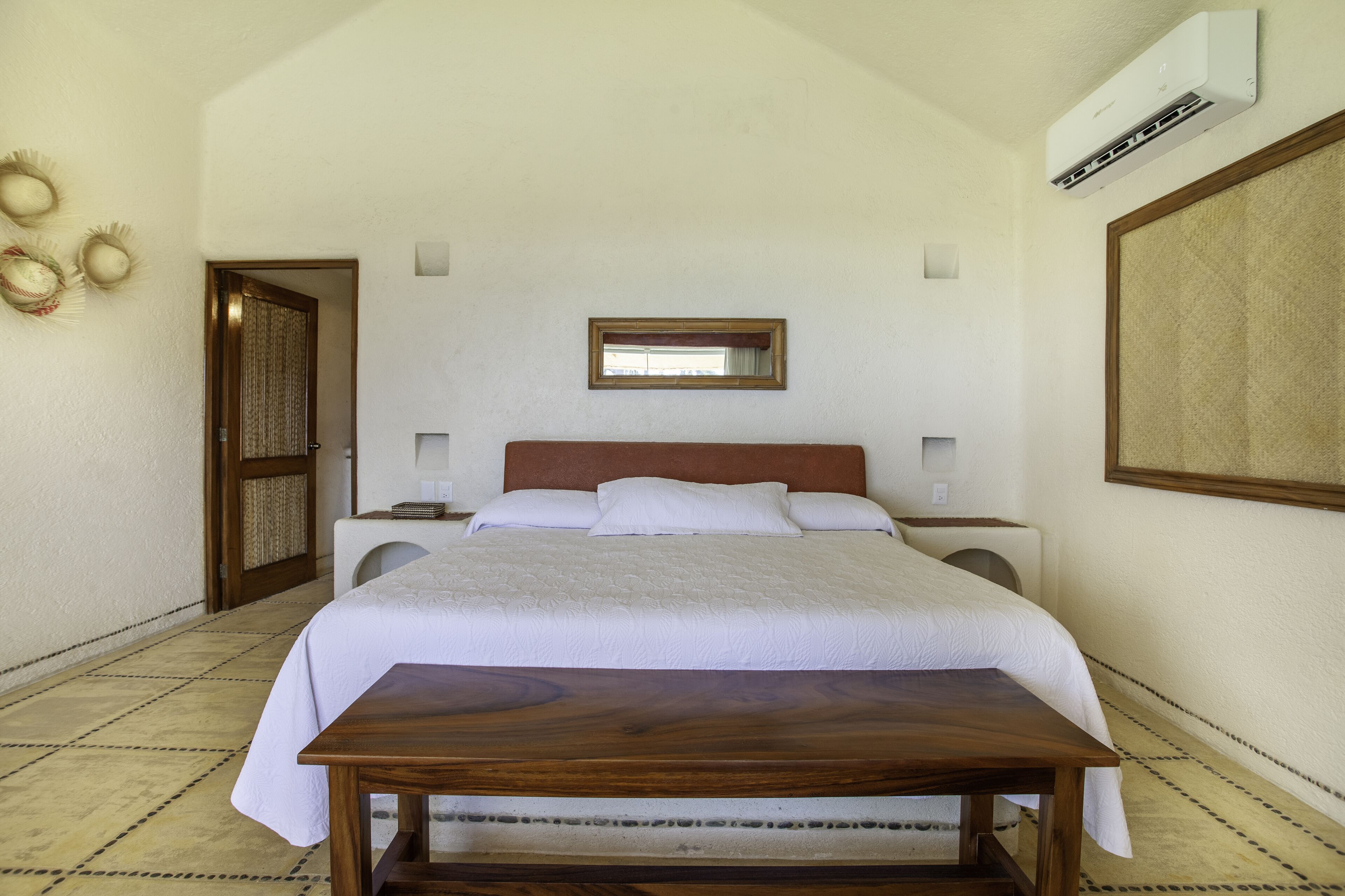 Quarto de hotel com cama, mesas de cabeceira, uma mesa de madeira, espelho e ar condicionado.