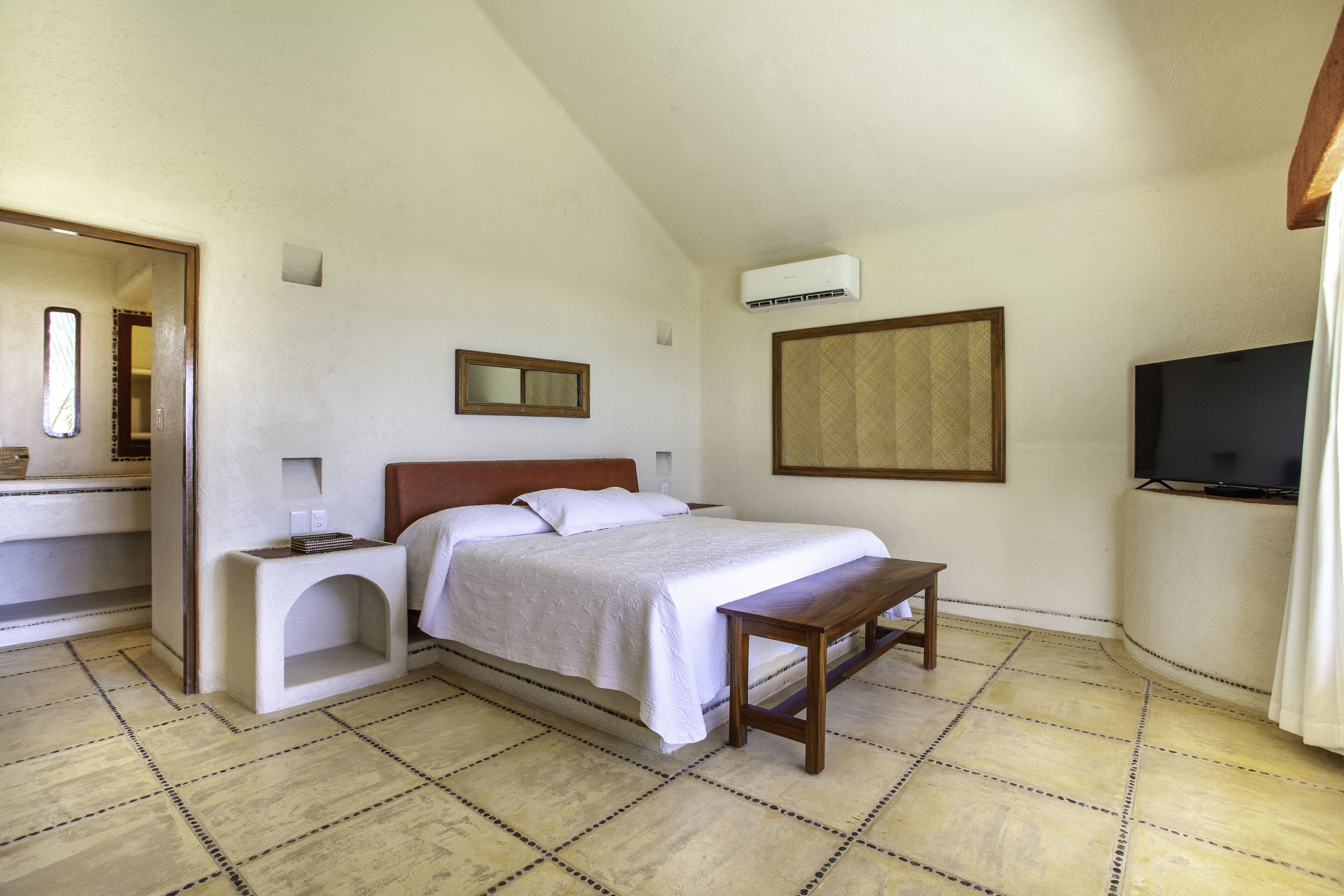 A bedroom with a bed, bedside table, mirror, air conditioning unit, television, and a bathroom visible through an open door.