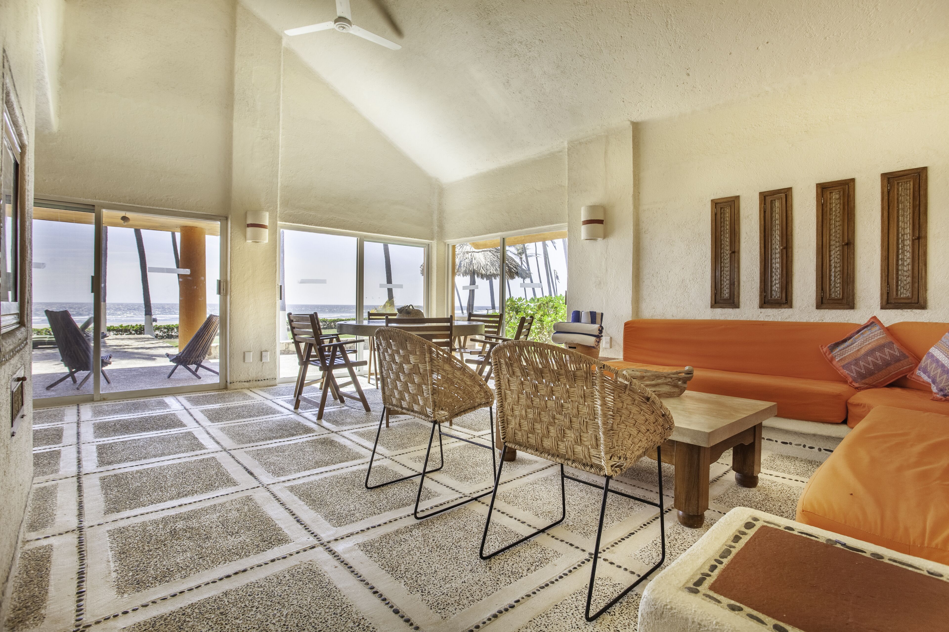 A spacious living area with a large window overlooking the sea, wicker chairs, an orange sofa, and a patterned rug.