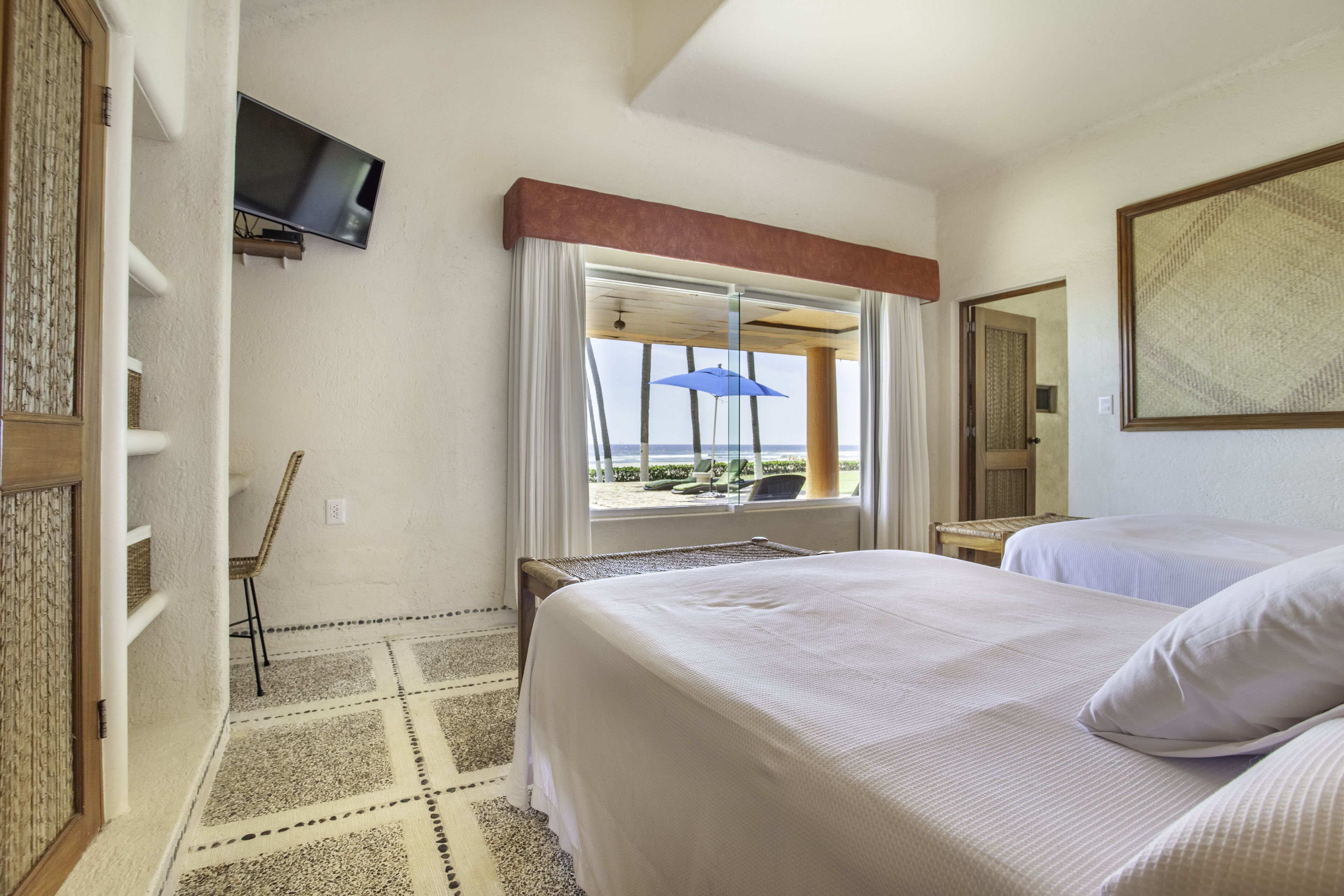 A hotel room with two beds, a TV mounted on the wall, a window with a view of the beach, and a patterned rug.