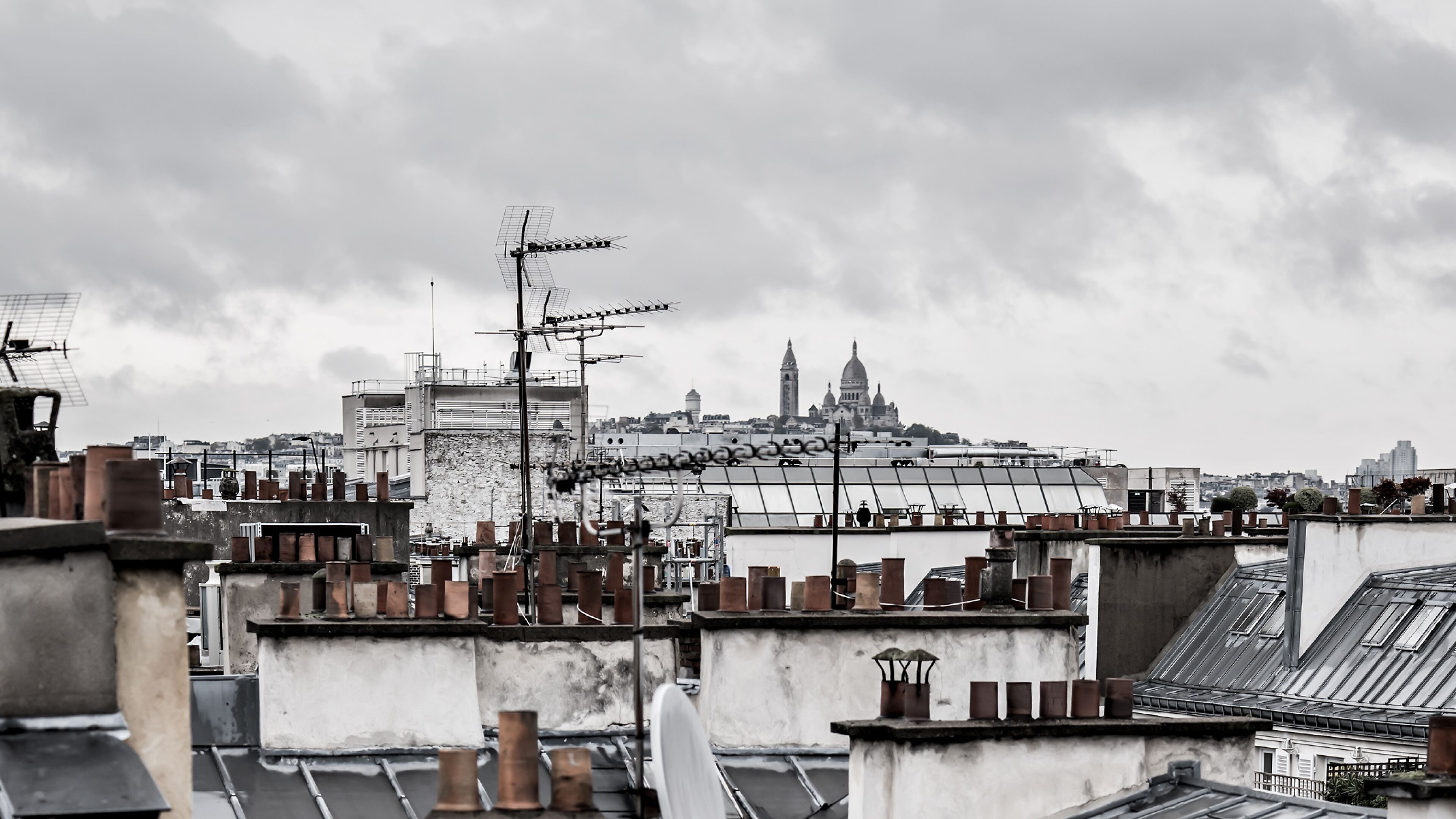 A rooftop with multiple chimneys and antennas, overlooking a cityscape with historical buildings.