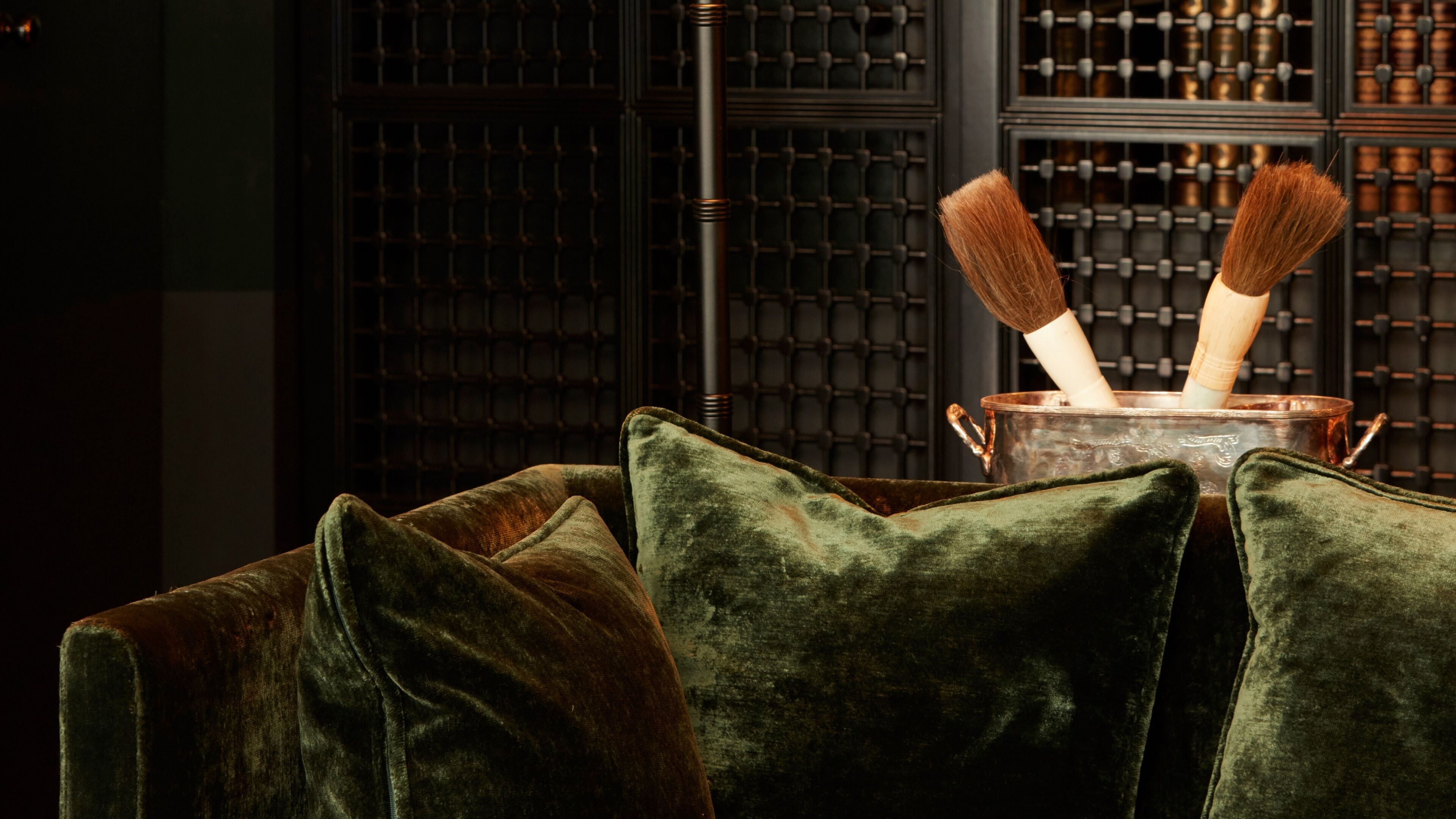 Una habitación con un sofá de terciopelo verde, una olla de cobre con asa de madera y un panel de pared de metal al fondo.