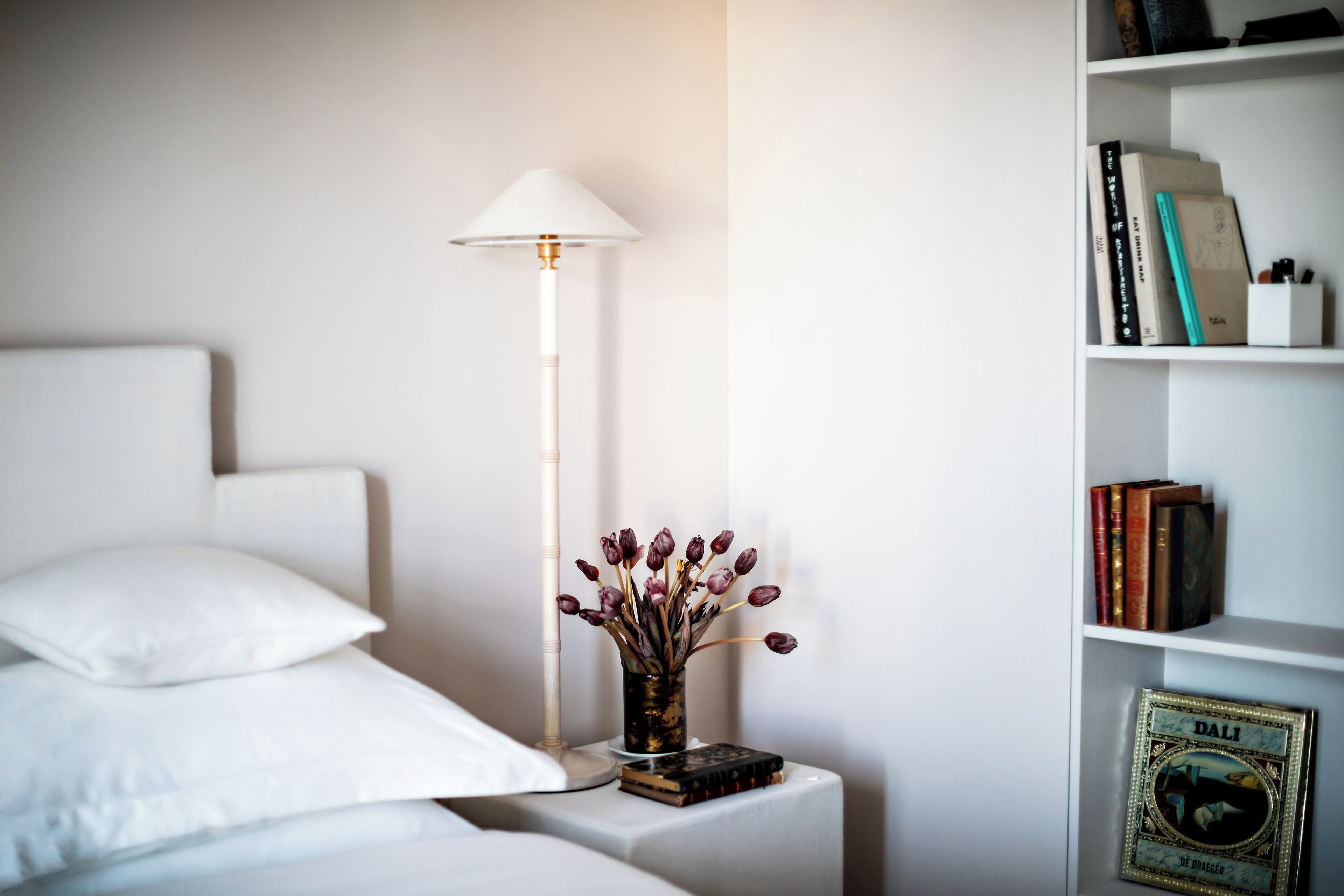 Un lit bien fait, avec du linge de lit blanc, une lampe de chevet, une petite table sur laquelle est posé un vase de fleurs et une bibliothèque remplie de livres et ornée d’un tableau encadré.
