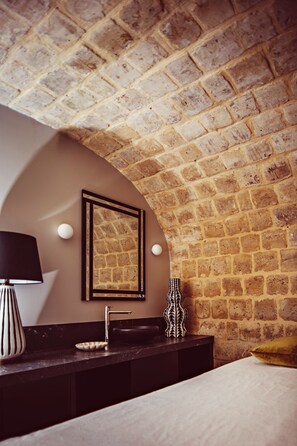 A bathroom with a stone wall, a framed mirror, a sink, a black lamp, and a decorative vase.