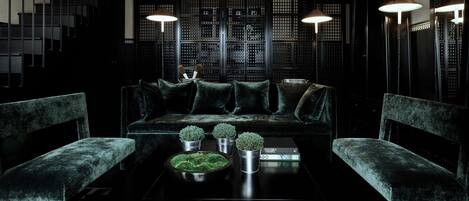 Un salón con decoración oscura, que incluye un sofá negro, dos sillones, una mesa de centro con plantas decorativas y un separador de ambientes de estilo asiático en el fondo.