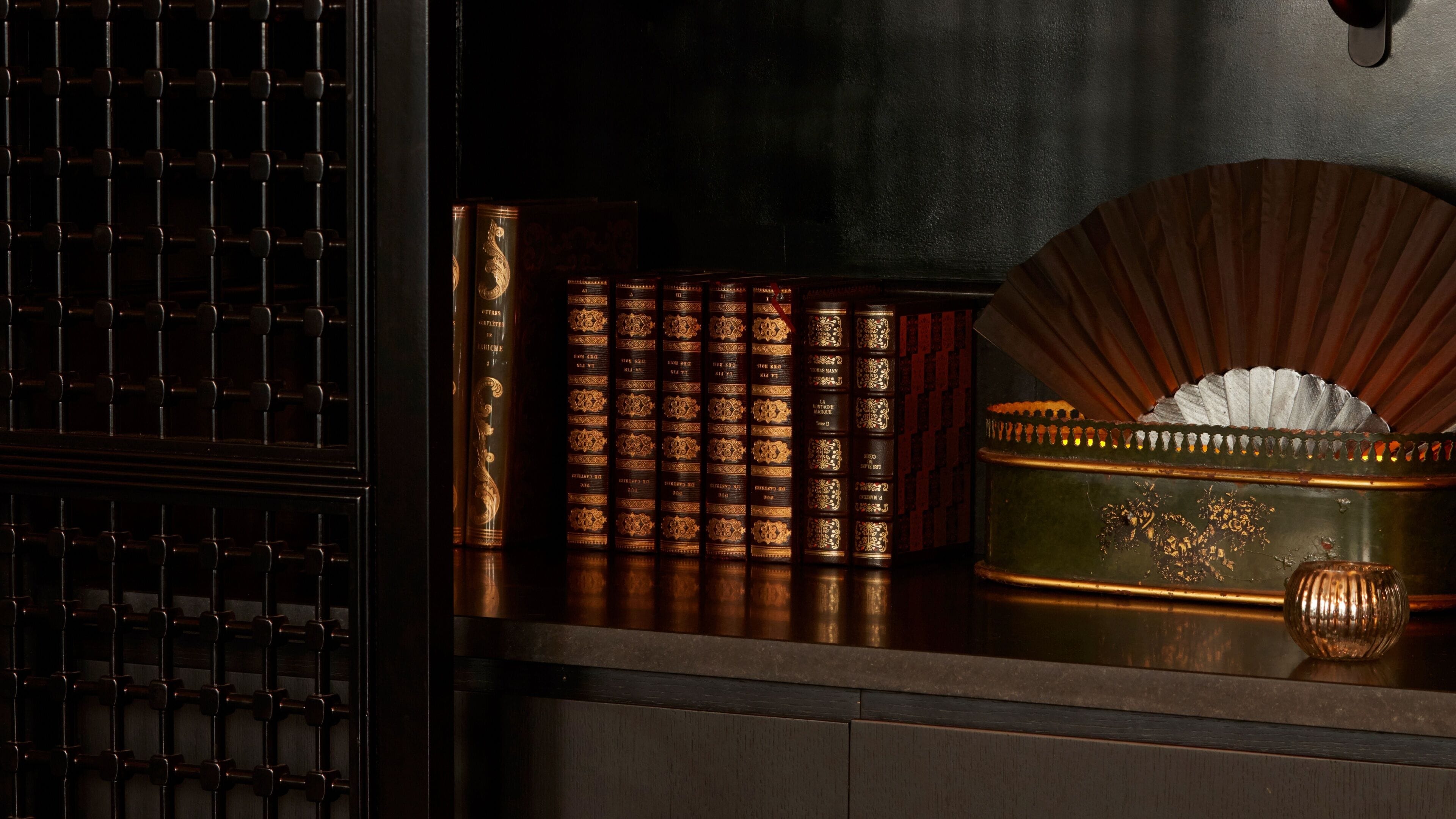 Un armario de madera con puerta de rejilla metálica, una pila de libros ornamentados y una caja verde con tapa decorativa.