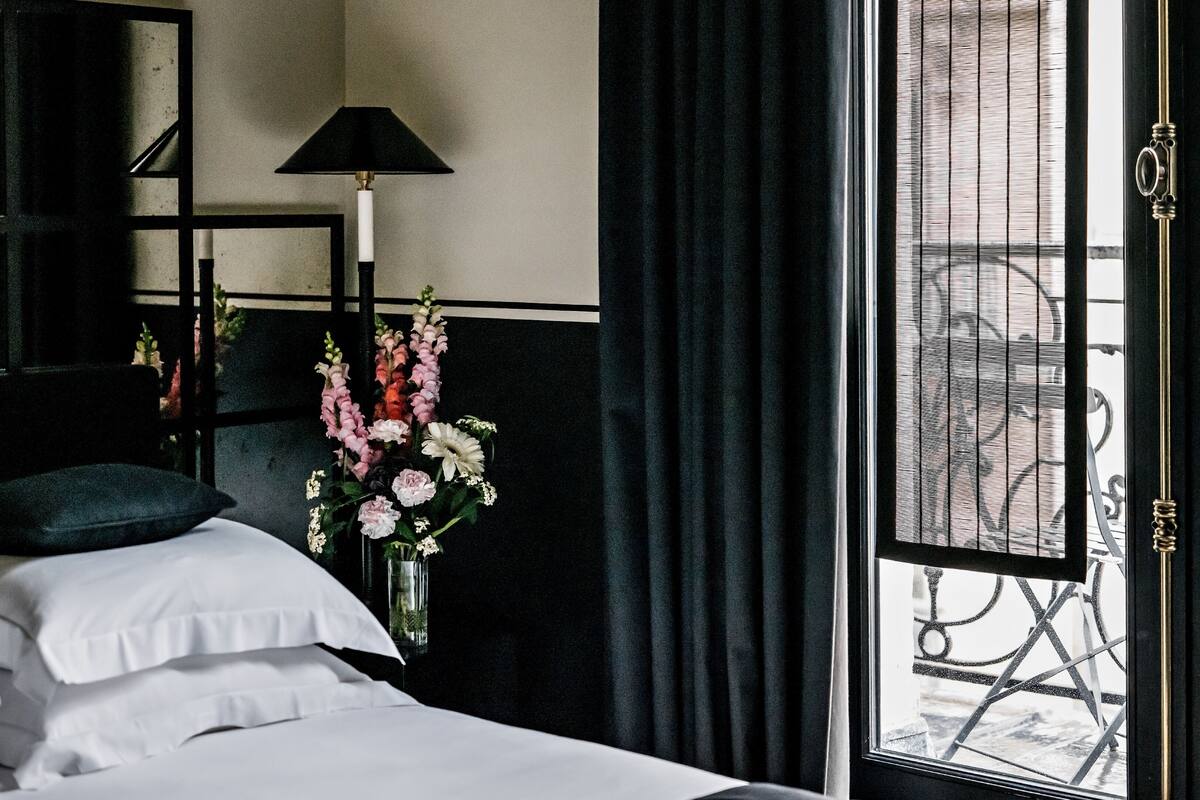 A hotel room with a bed, pillows, a vase with flowers, a lamp, and a window with curtains.