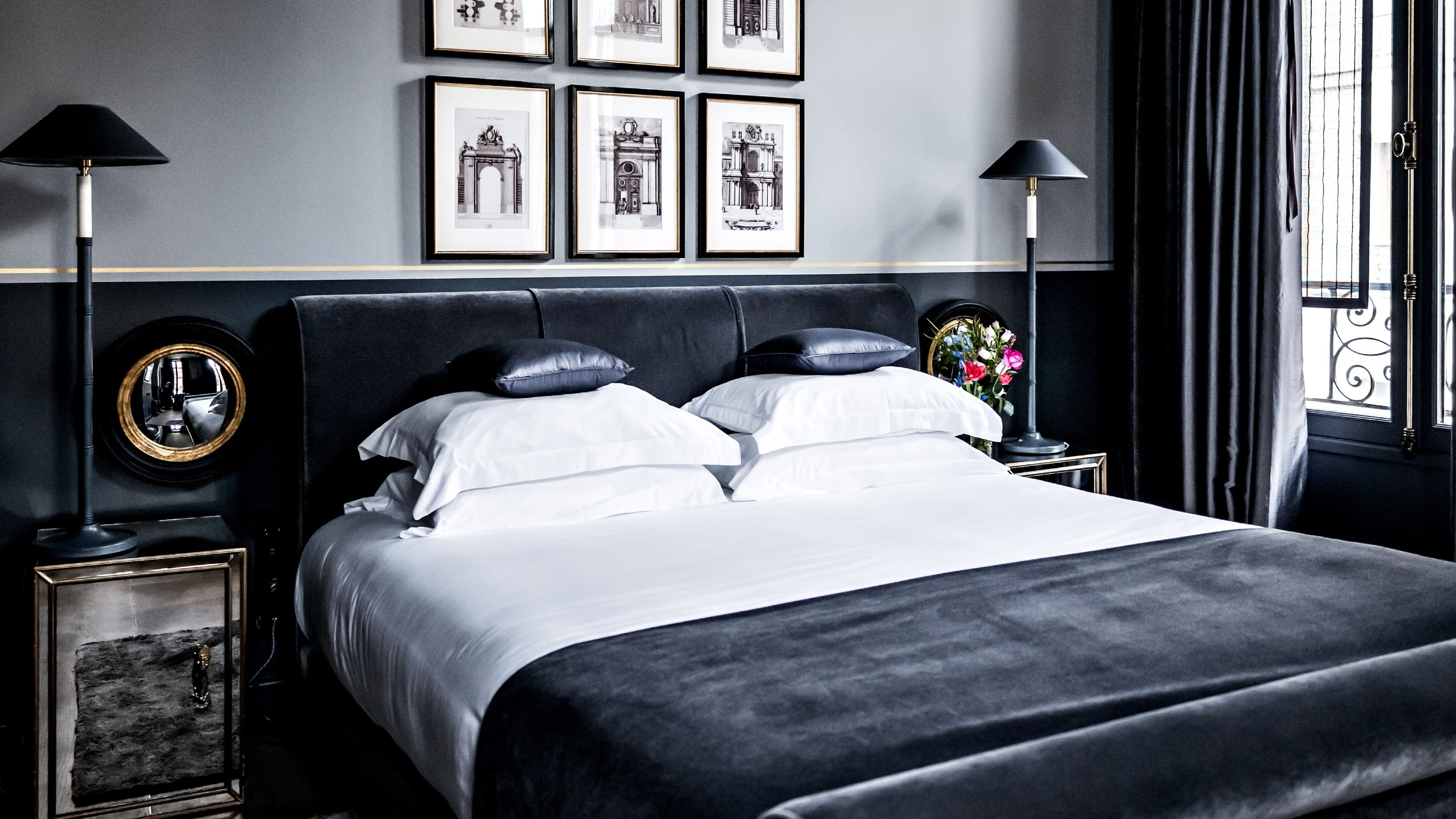A neatly made bed with white linens and black pillows, a round mirror, and a framed picture on a nightstand.