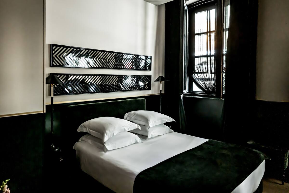 A modern hotel room with a large bed, a decorative headboard, and a window with blinds.