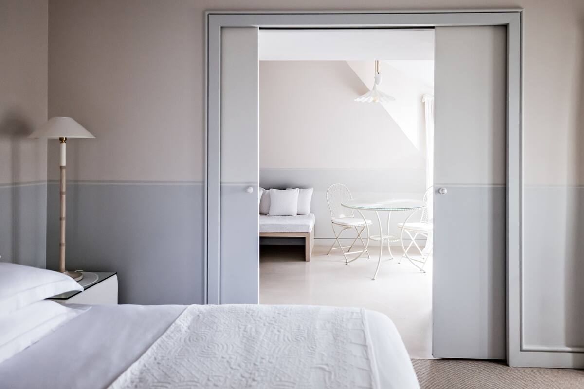 A hotel room with a bed, a nightstand, a lamp, a dining area with a table and chairs, and a bathroom visible through an open door.