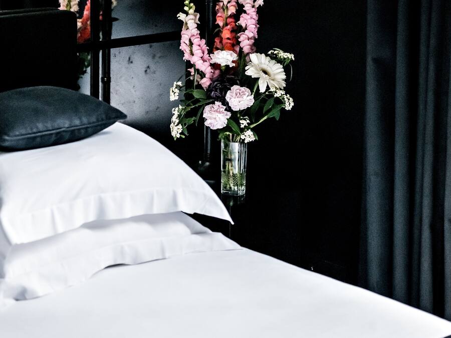 A neatly made bed with white linens and a dark pillow, a vase with flowers on a bedside table, and a dark curtain in the background.