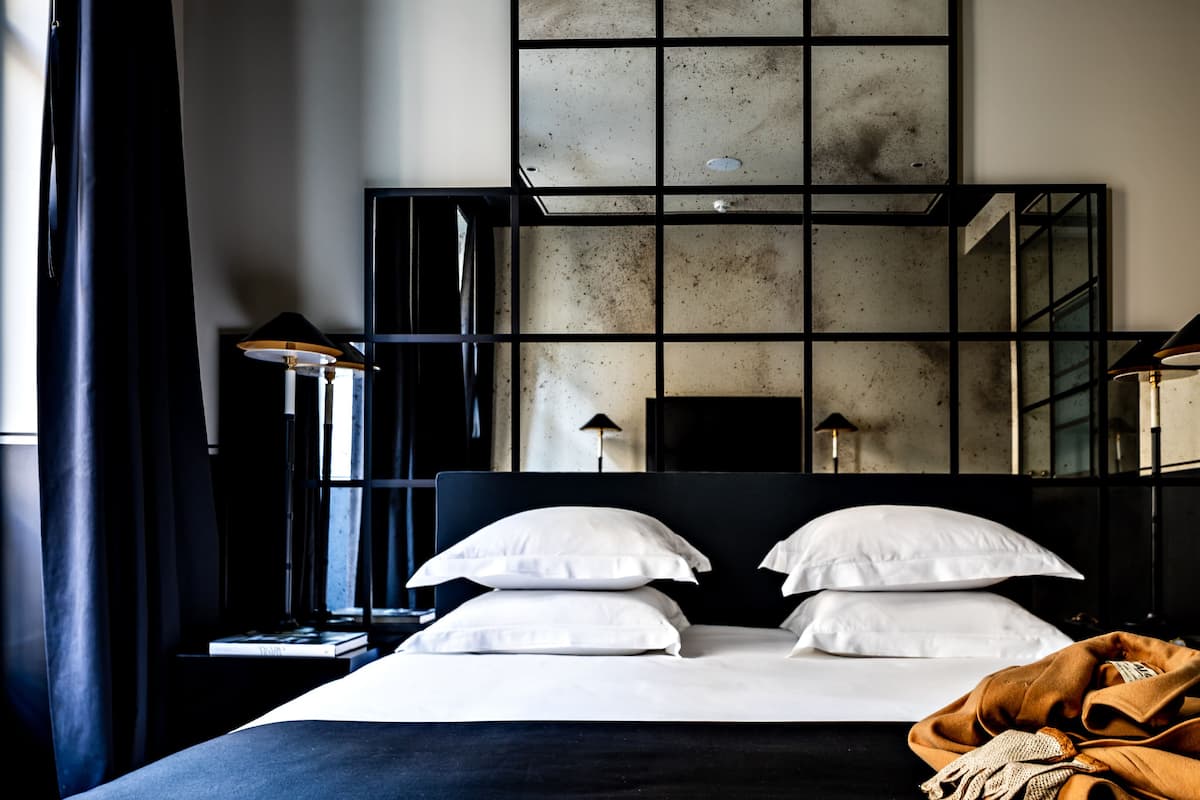 A modern hotel room with a large bed, two bedside lamps, a large mirror, and a view of the cityscape through the window.